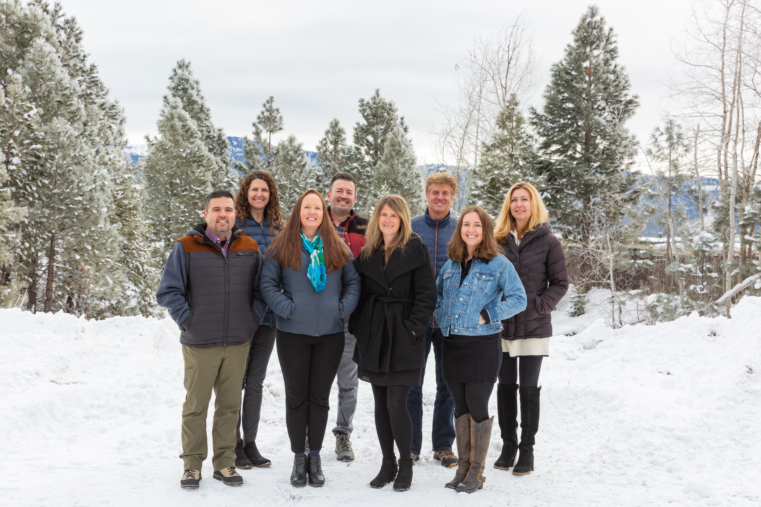 Eight people standing together in the snow with frosted pine trees behind them. 
