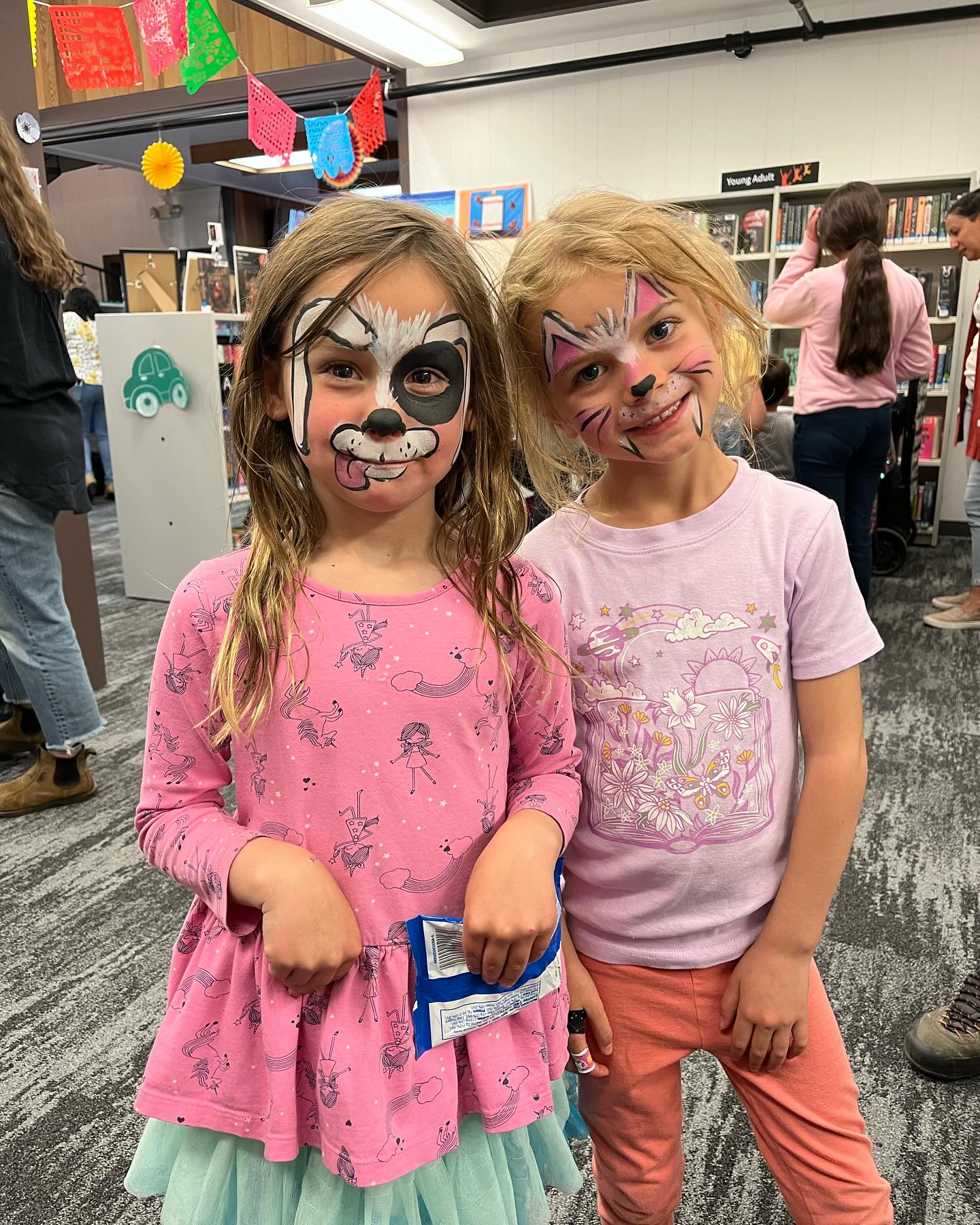 Two children with faces painted smiling together in a library. 