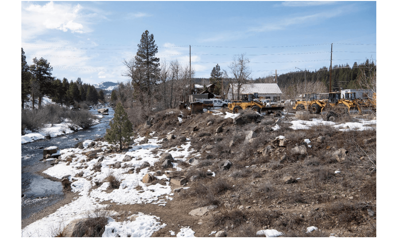A photo of the Truckee River with snow on banks, dirt and rocks up the hill to row of tractors. 